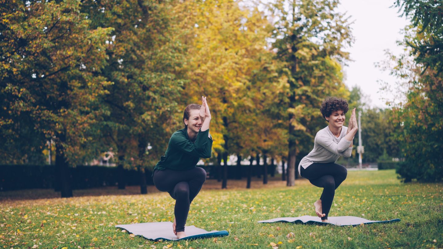 Two women doing yoga on mats in an autumn park, surrounded by colorful trees.
