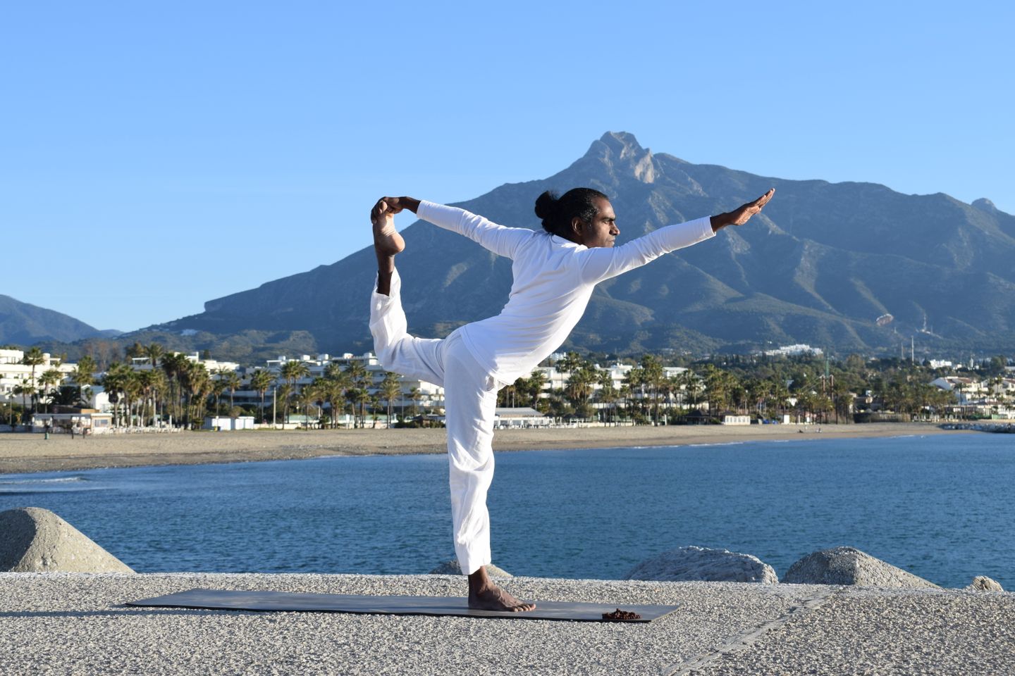 Man practicing yoga pose on beach with mountains in background.